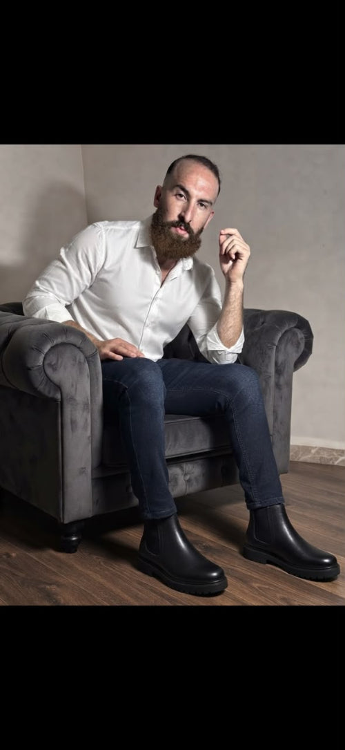 A man with a full beard sits in a gray armchair wearing a white button-down shirt, dark blue jeans, and black leather ankle boots. The photo is taken in an indoor setting with a neutral beige wall and wood floor, framed with black letterbox bars at the top and bottom.