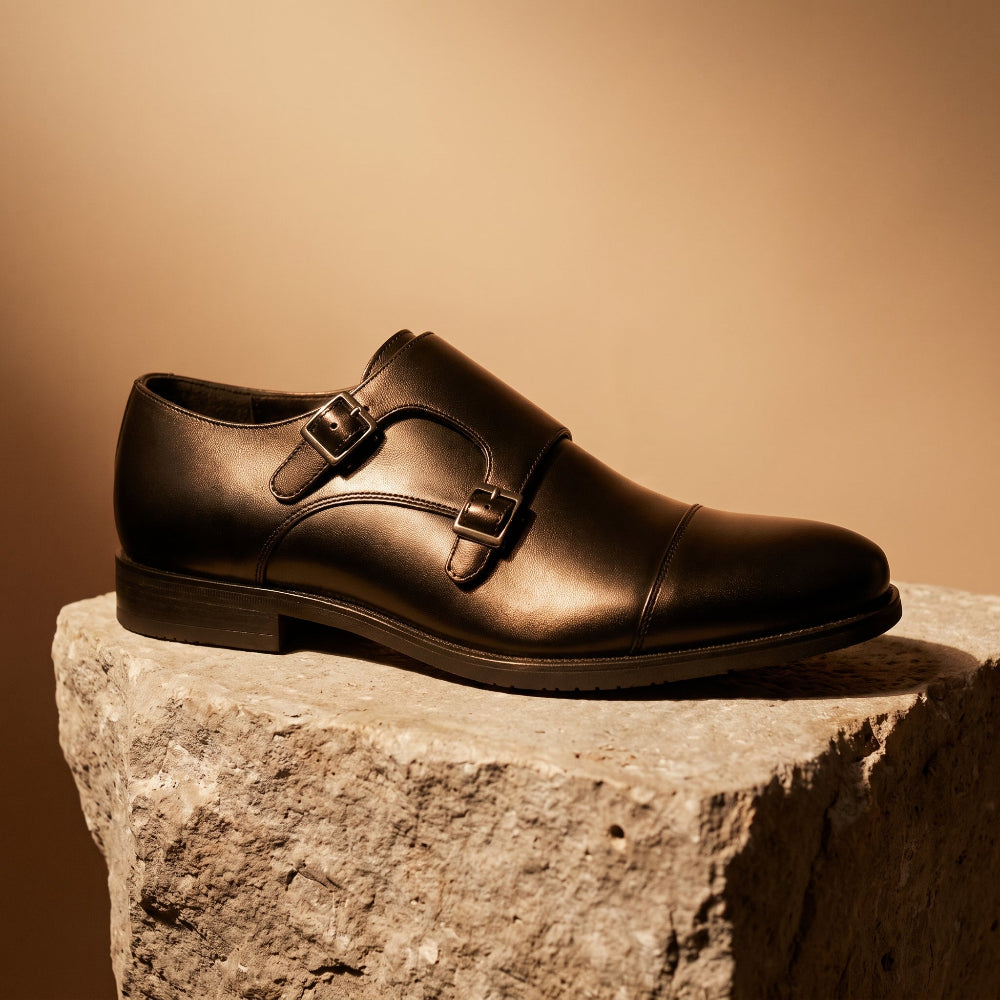 A black leather double strap monk-style dress shoe with a cap toe, displayed on a rough stone pedestal against a warm beige background.