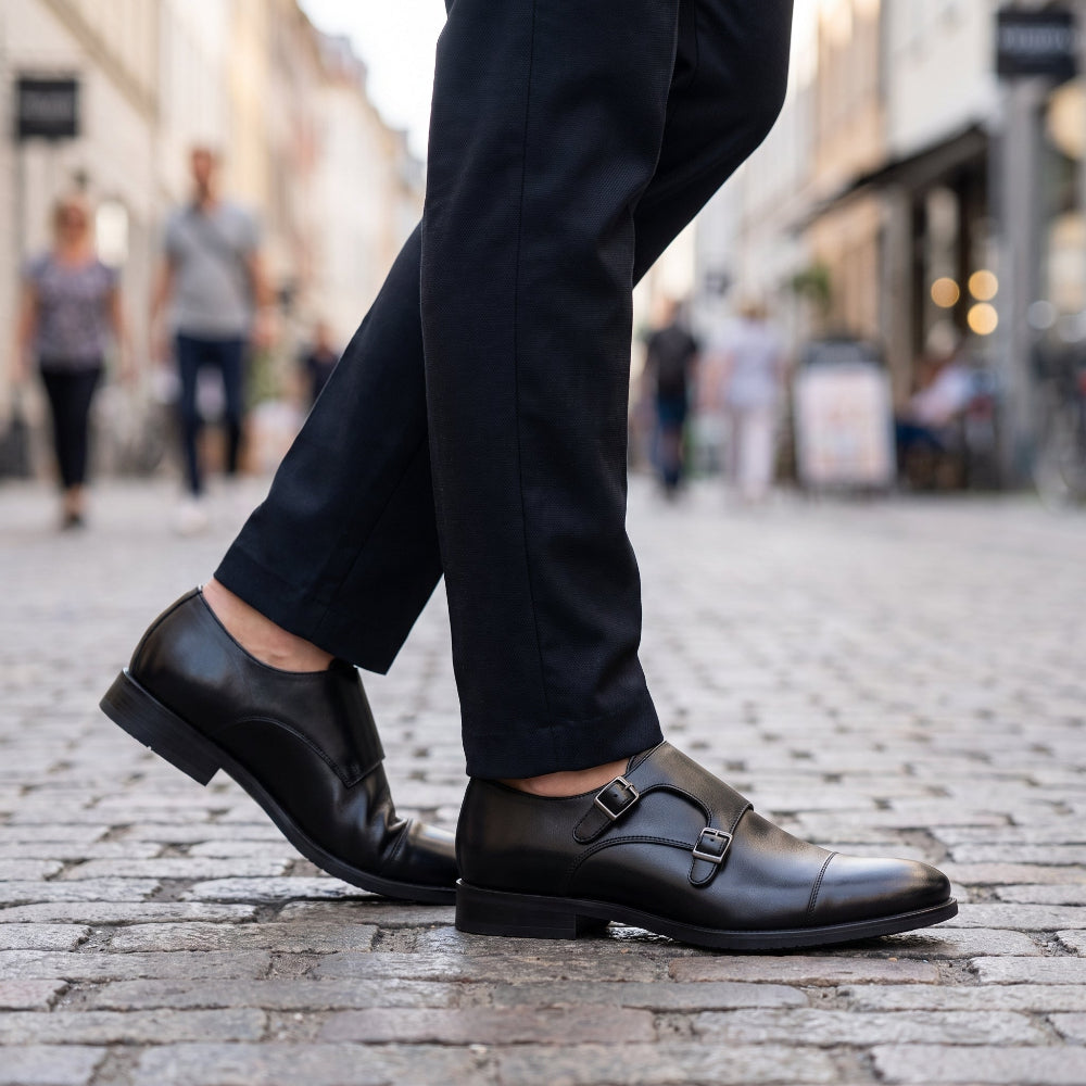 A close-up of a person's legs wearing black double monk strap leather dress shoes and dark trousers, walking on a cobblestone street with a blurred city background.