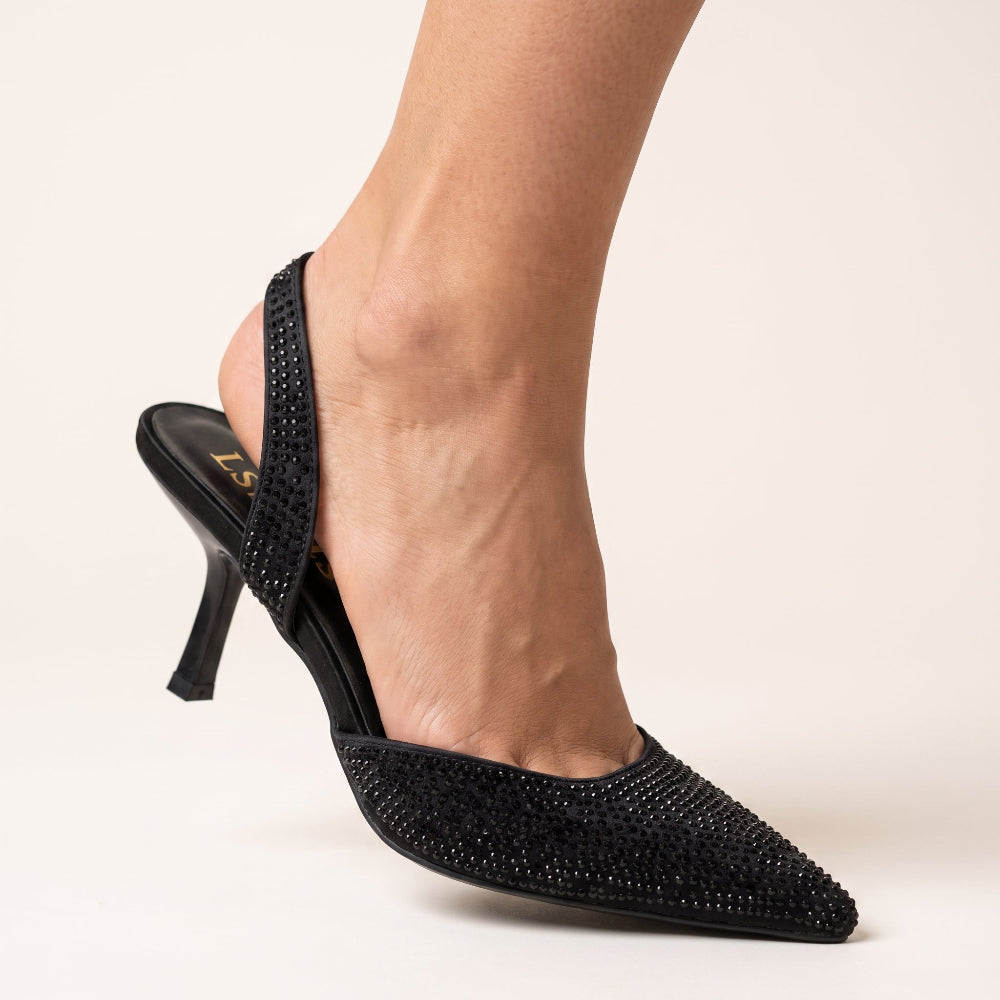 A close-up of a person's foot wearing a black pointed-toe slingback high heel shoe embellished with sparkling rhinestones, set against a plain light beige background.