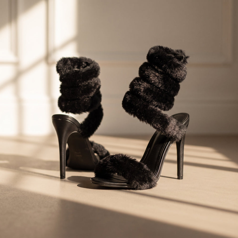 A pair of black high-heeled sandals with thick, fluffy faux-fur straps wrapping around the foot and ankle, photographed in warm sunlight on a beige floor.