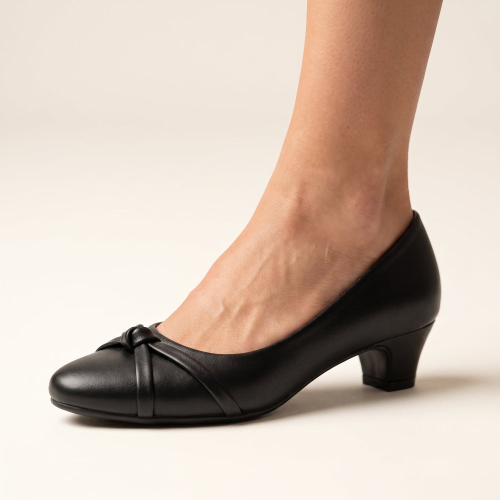 A woman's foot wearing a brown leather pump shoe with a low block heel and a small bow detail on the toe, shown against a plain light background.