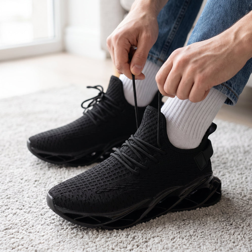 A person wearing black knit athletic sneakers with white socks, sitting on a light carpet and tying the laces of the front shoe.