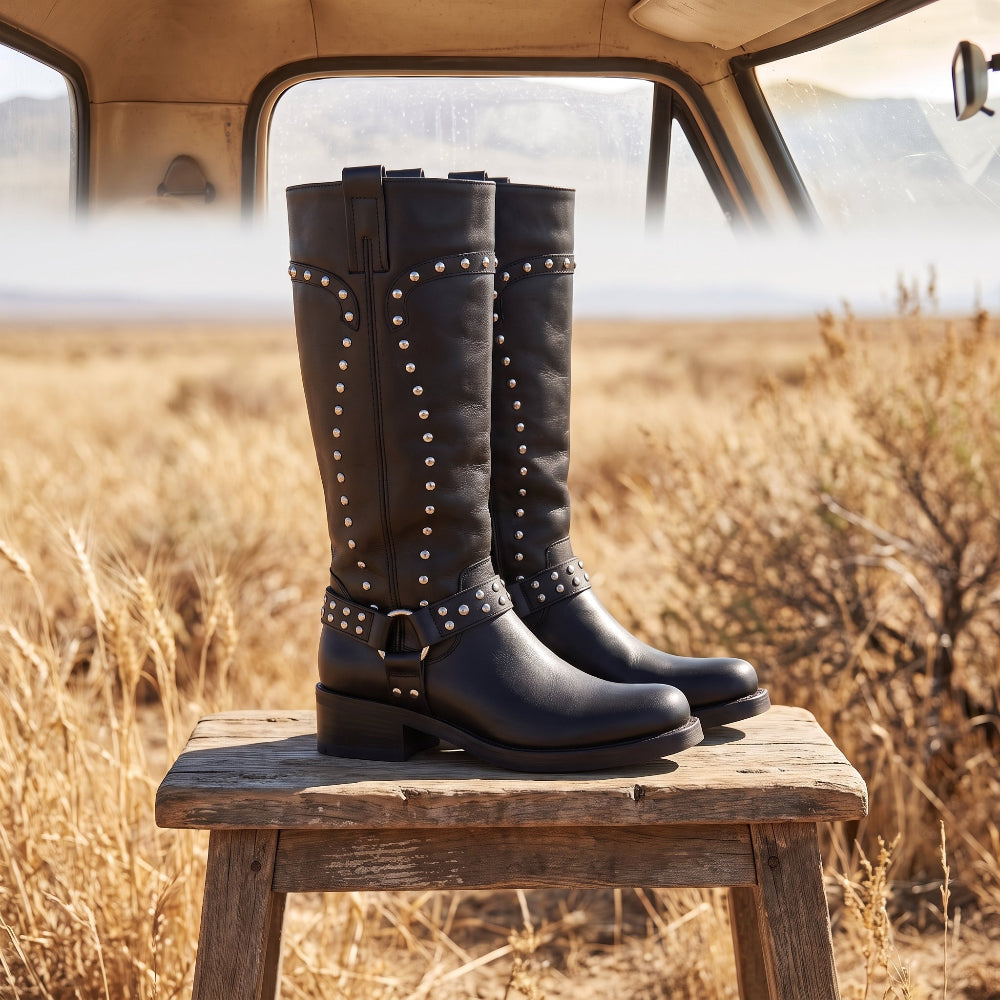 A pair of tall black leather boots with silver studs and buckle details, displayed on a rustic wooden stool in an outdoor field with a vintage car interior in the background.
