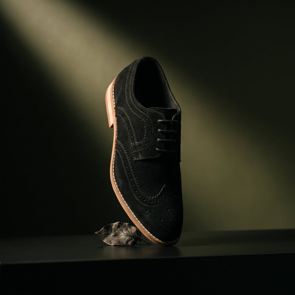 A single black suede wingtip oxford shoe with brogue detailing and black laces, standing on a dark surface with a small gray rock prop. Dramatic spotlighting creates a warm glow on the background.