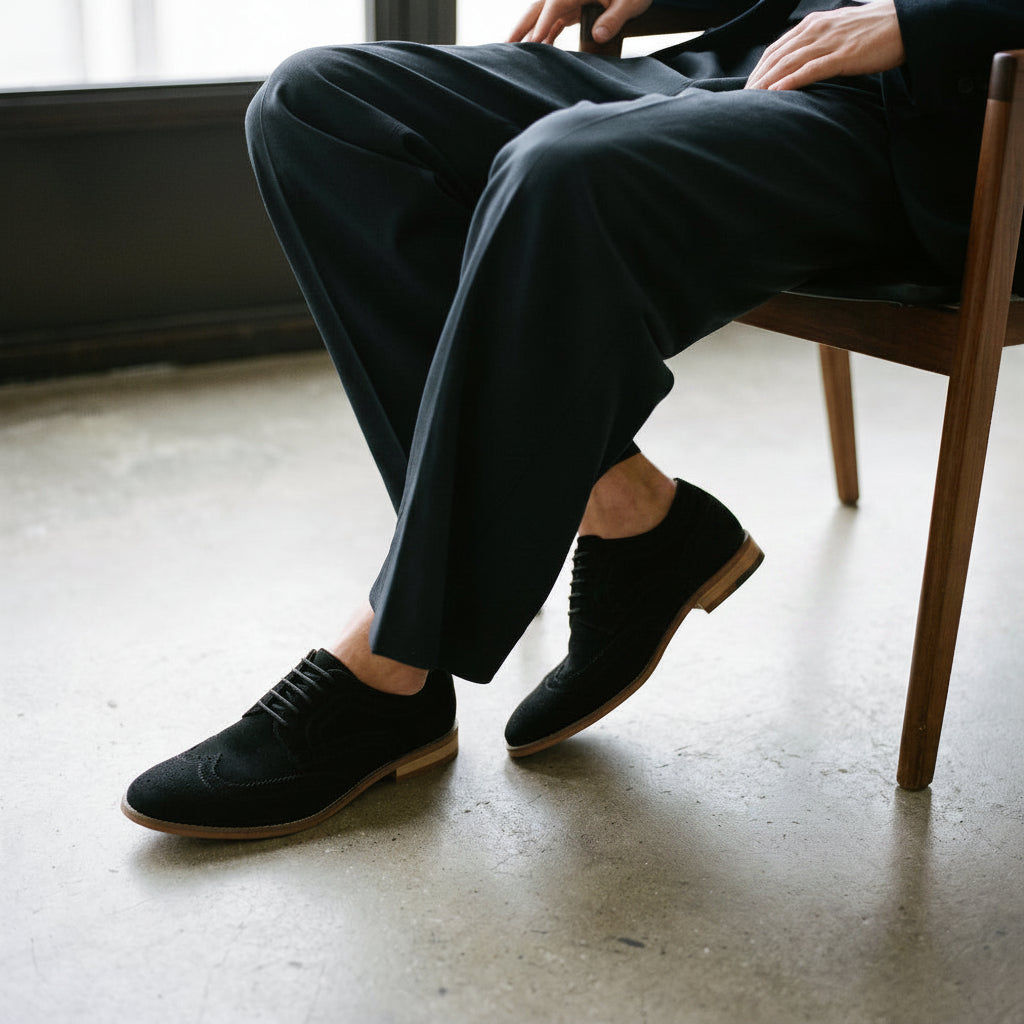 A person seated on a wooden chair wearing black suede oxford-style shoes with brogue detailing and dark trousers, photographed in a real indoor setting with a concrete floor and window light.