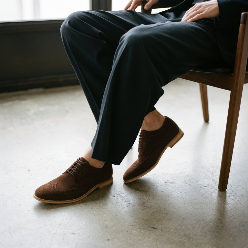 A person seated on a wooden chair wearing black suede oxford-style shoes with brogue detailing and dark trousers, photographed in a real indoor setting with a concrete floor and window light.