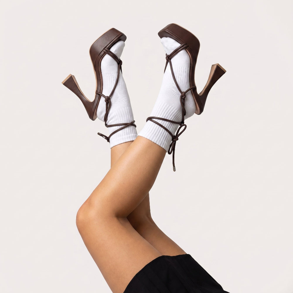 A pair of brown strappy high-heeled sandals with chunky block heels, styled with white ribbed crew socks on crossed legs against a light beige studio background.