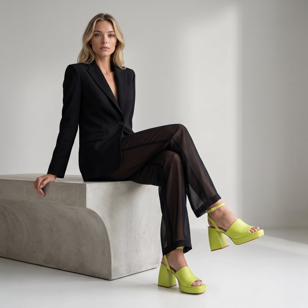 A woman sits on a concrete bench wearing a black tailored blazer and sheer black wide-leg pants, paired with bright neon yellow platform sandals, against a clean light gray studio background.