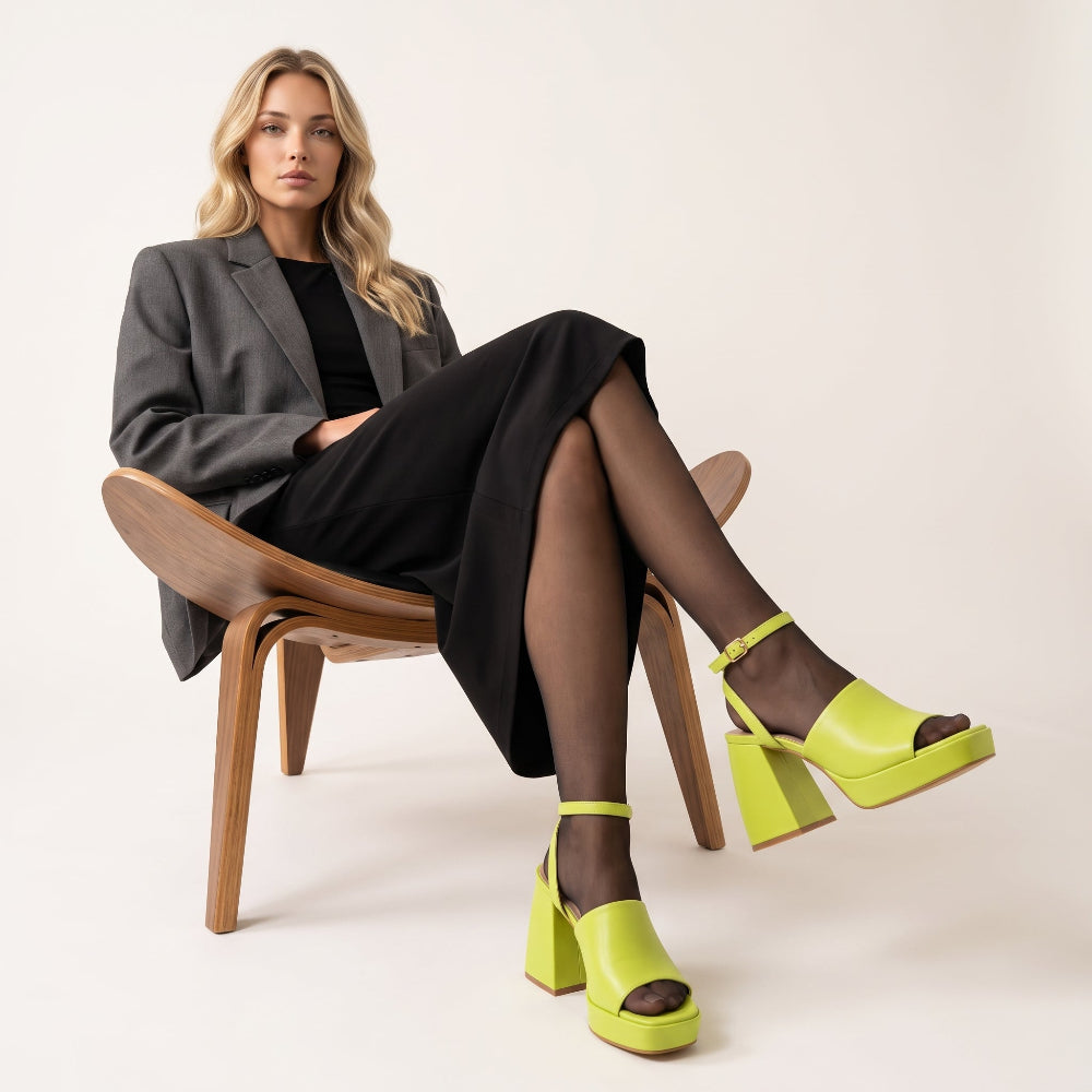 A woman sits on a wooden chair wearing a gray blazer, black skirt, black tights, and bright neon yellow platform sandals with ankle straps and block heels against a plain light beige studio background.