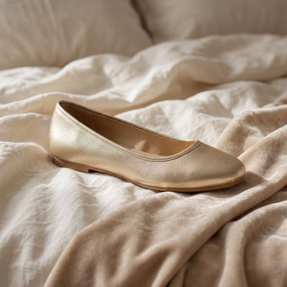 A single metallic champagne flat shoe resting on rumpled beige fabric, with a soft neutral background.