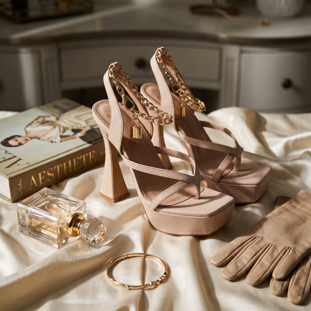 A pair of beige slingback high-heeled sandals with gold chain and padlock ankle details, styled on draped cream satin with a book, perfume bottle, bangle, and gloves nearby.