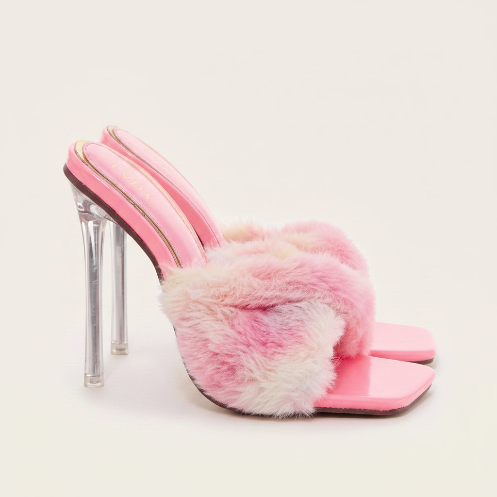 A pink patent leather mule sandal with a fluffy pink and white faux fur strap, a clear high-heel stiletto, and gold trim on a light beige background.