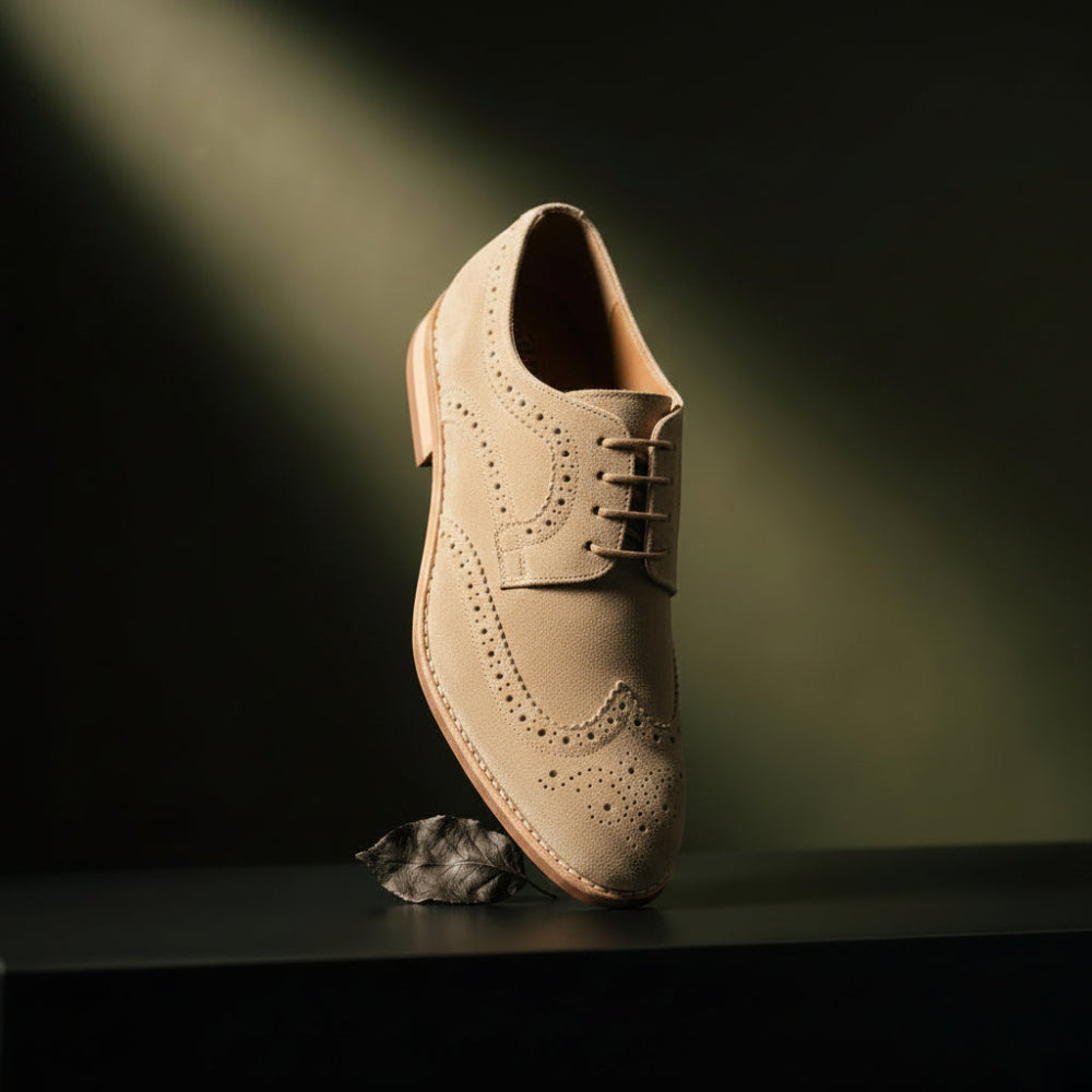 A singleTAN suede wingtip oxford shoe with brogue detailing and black laces, standing upright on a dark surface with a small leaf prop against a dark background with dramatic spotlighting.