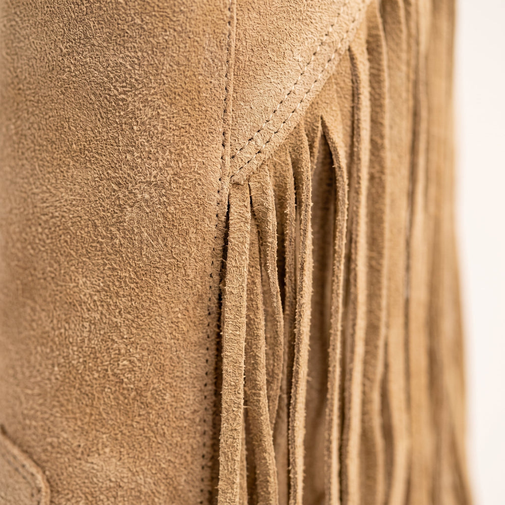 Close-up detail of a tan suede fringe garment, showing the soft suede texture and hanging fringe against a light neutral background.