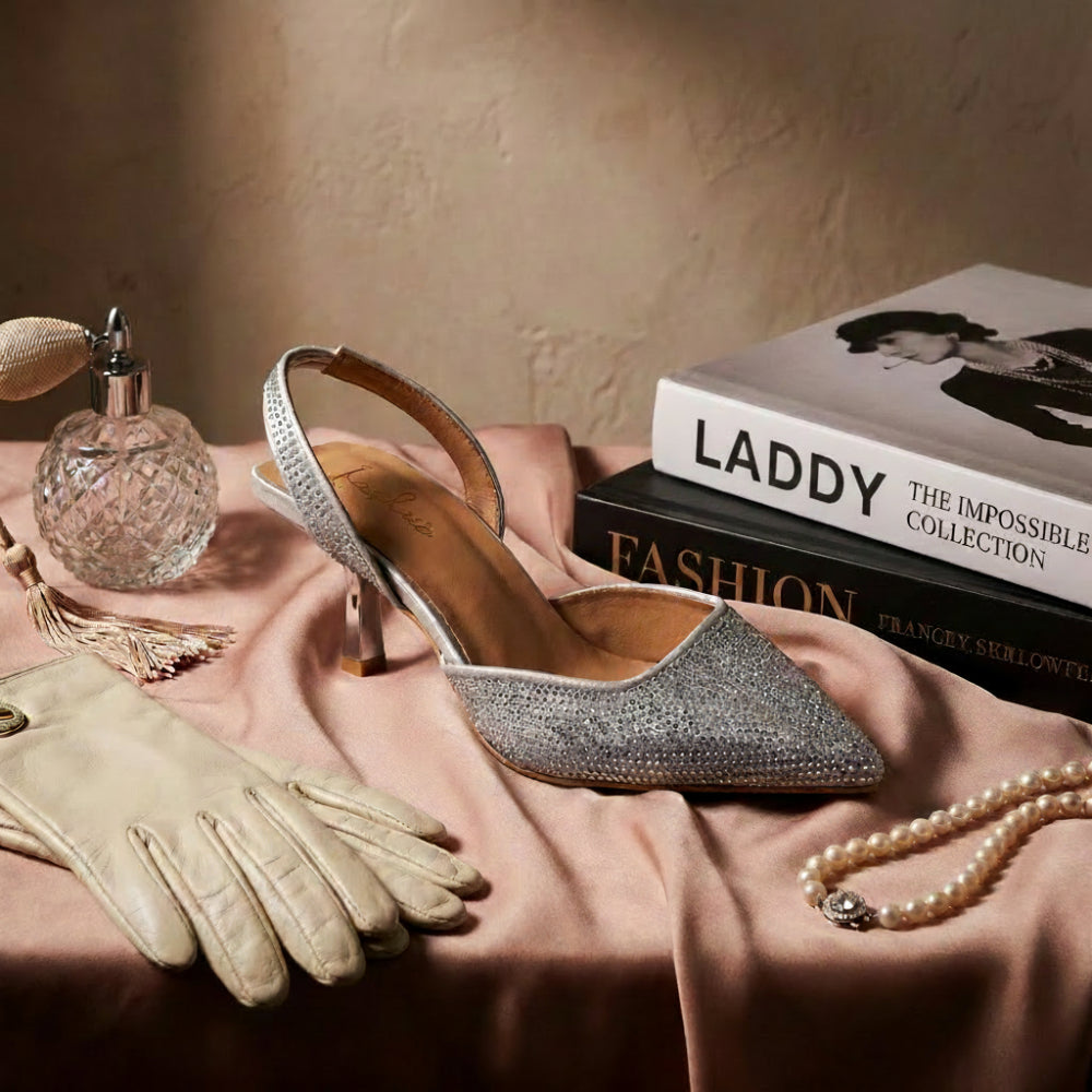 A single pointed-toe slingback high heel in sparkling silver rhinestones, styled on draped blush pink satin with a cream glove, perfume bottle, pearl necklace, and a stack of fashion books including one labeled Chanel.